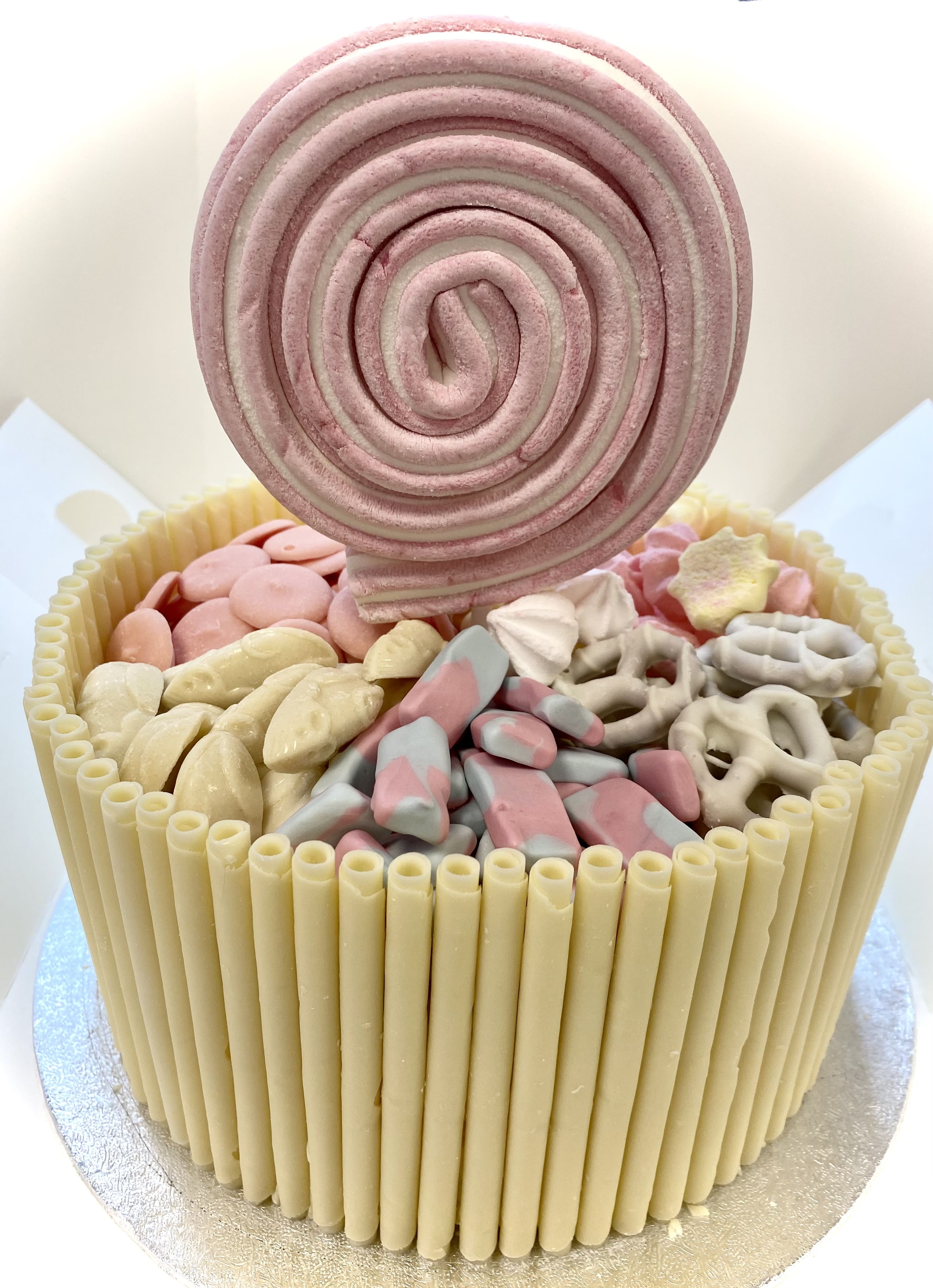 A cake adorned with vertical white chocolate sticks around its side, topped with various candies in shades of pink, white, and blue, including pretzels and marshmallows. A large pink spiral candy stands upright in the center as a decorative topper.